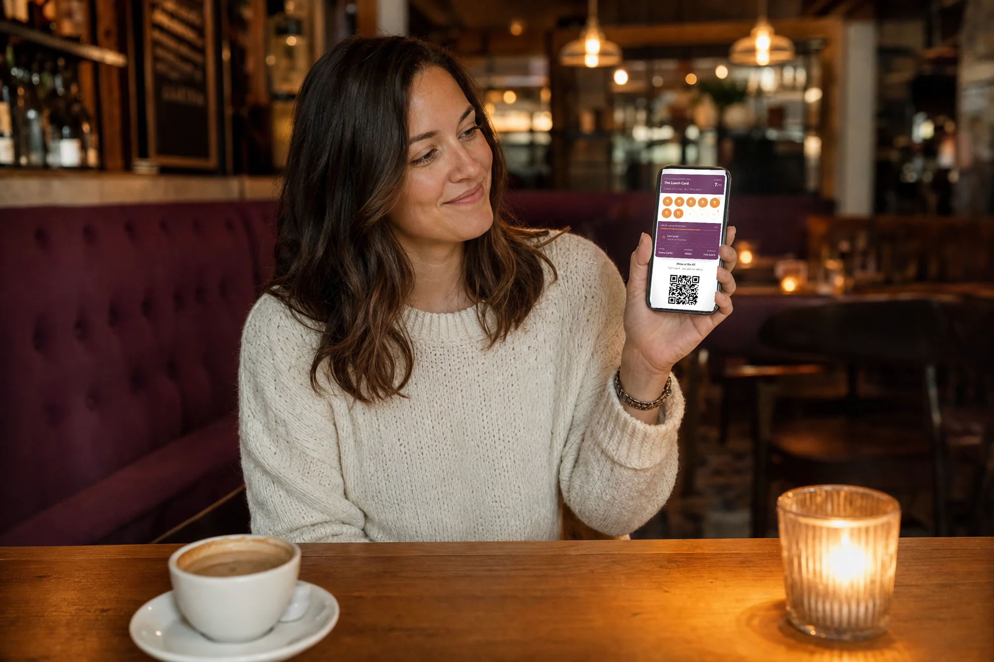 Guest at a table in a warm bistro showing the digital stamp card on their phone — seven of ten stamps in the lunch card and a QR code to scan at the till