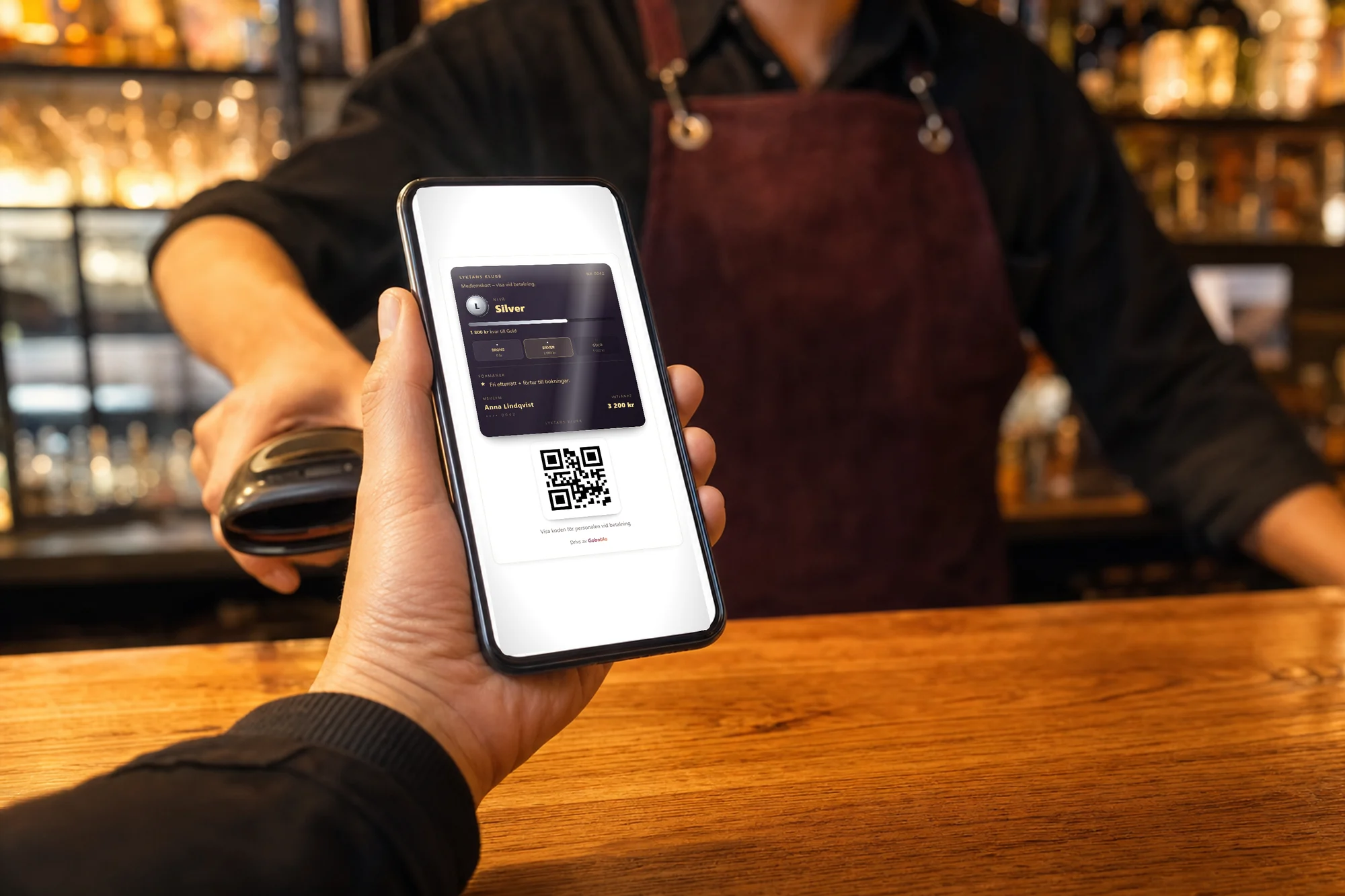 Gästens hand visar medlemskortet i mobilen vid kassan — Lyktans Klubb på nivå Silver med QR-kod, personal med skanner redo i bakgrunden