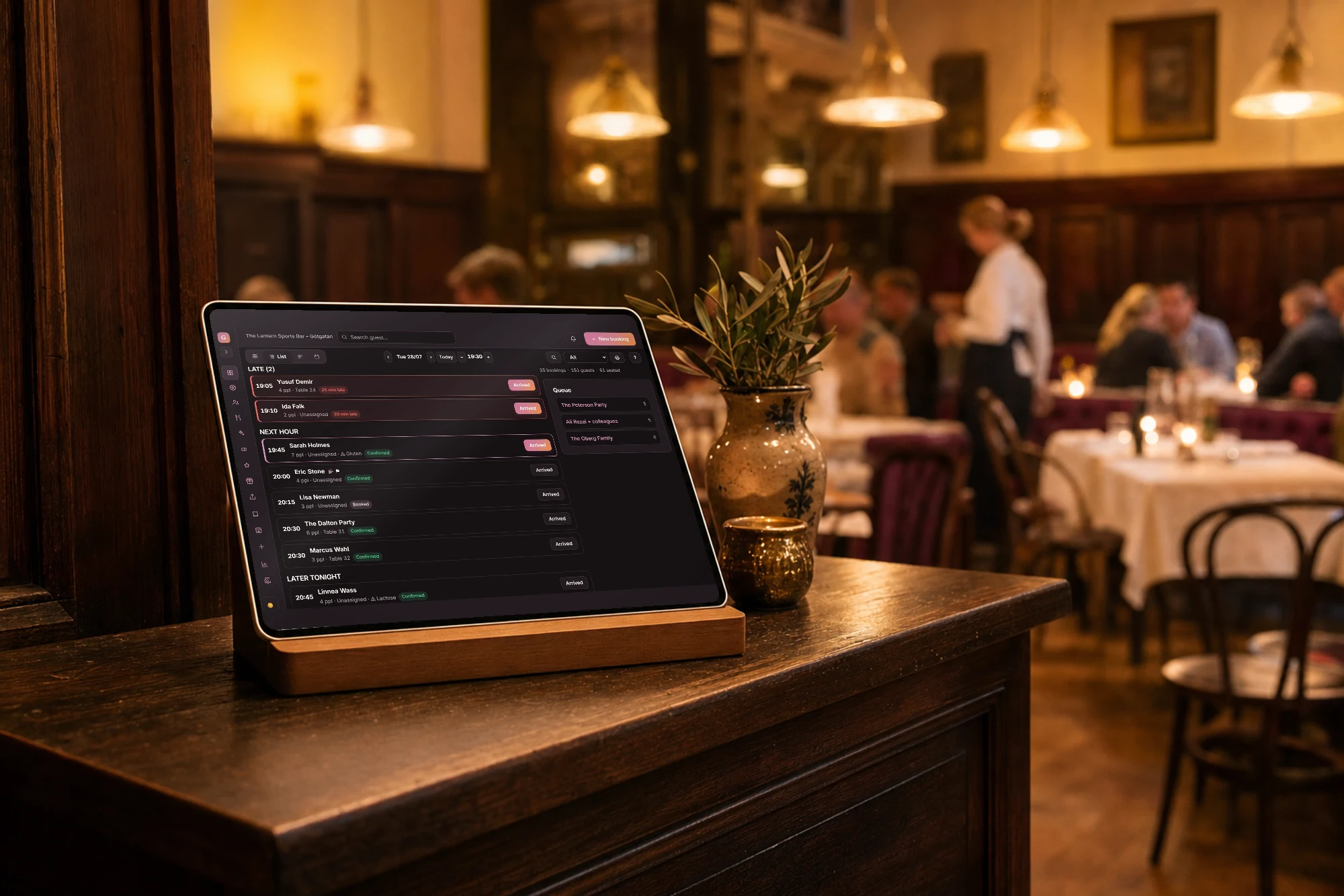 Empfangstresen eines Wiener Beisls im Abendservice: auf einem Tablet im Holzständer steht die Reservierungsliste des Abends in Goboblo – verspätete Gäste oben, Warteliste rechts – während der volle Gastraum dahinter unscharf bleibt