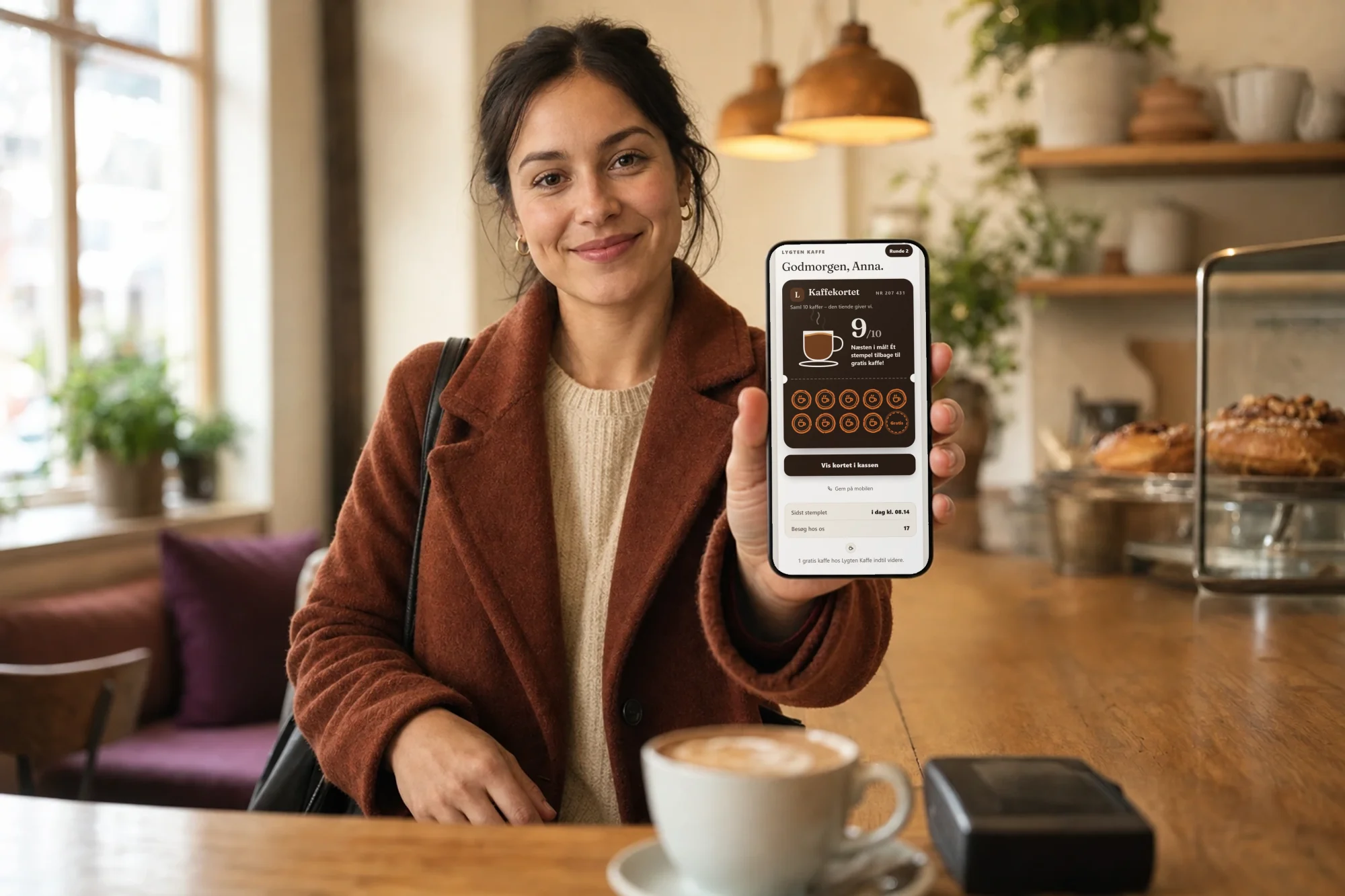 Gæst på en café holder mobilen op mod kassen. Skærmen viser stempelkortet til kaffe med 9 af 10 stempler og teksten »Næsten i mål! Ét stempel tilbage til gratis kaffe!«